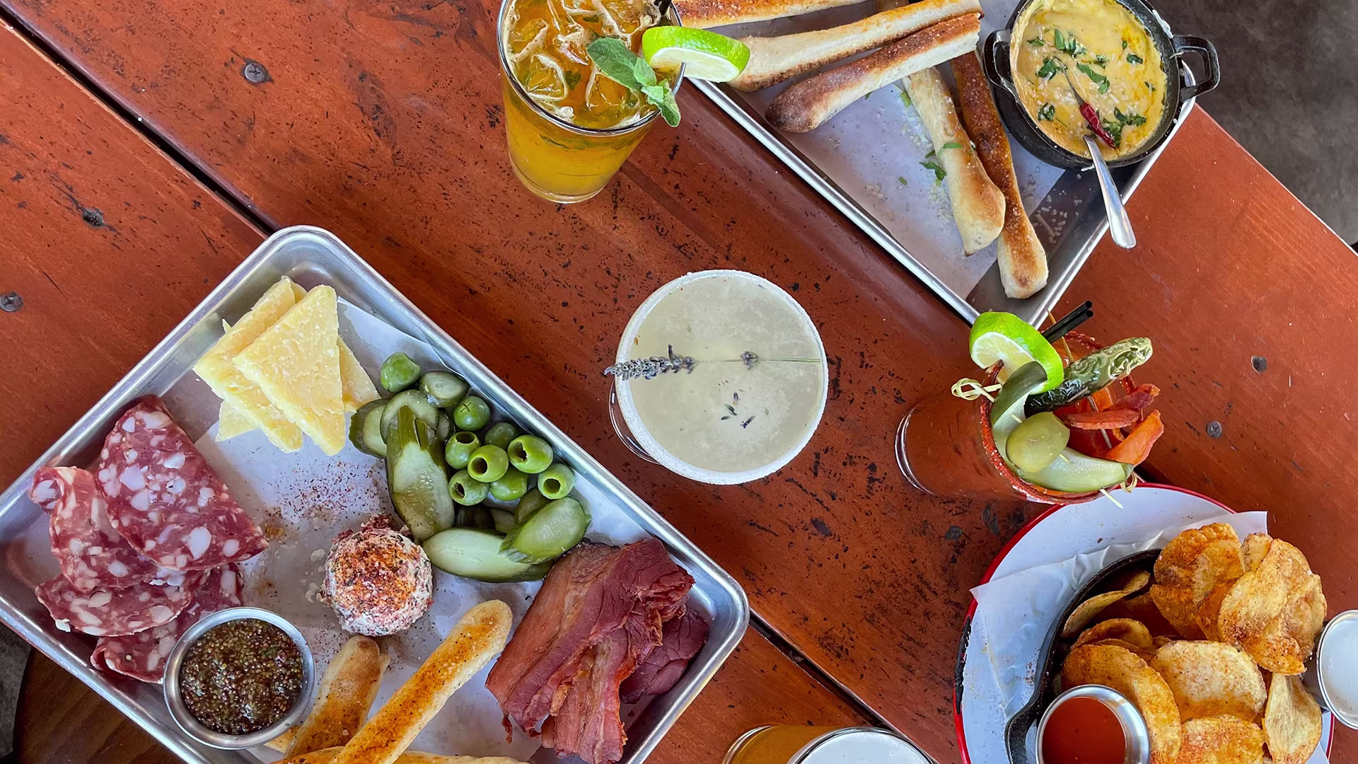 Table with charcuterie tray of meats, cheese, olives, pickles, mustard, breadsticks, drinks, chips with dipping sauce, and a Bloody Mary garnished with pickles and lime.
